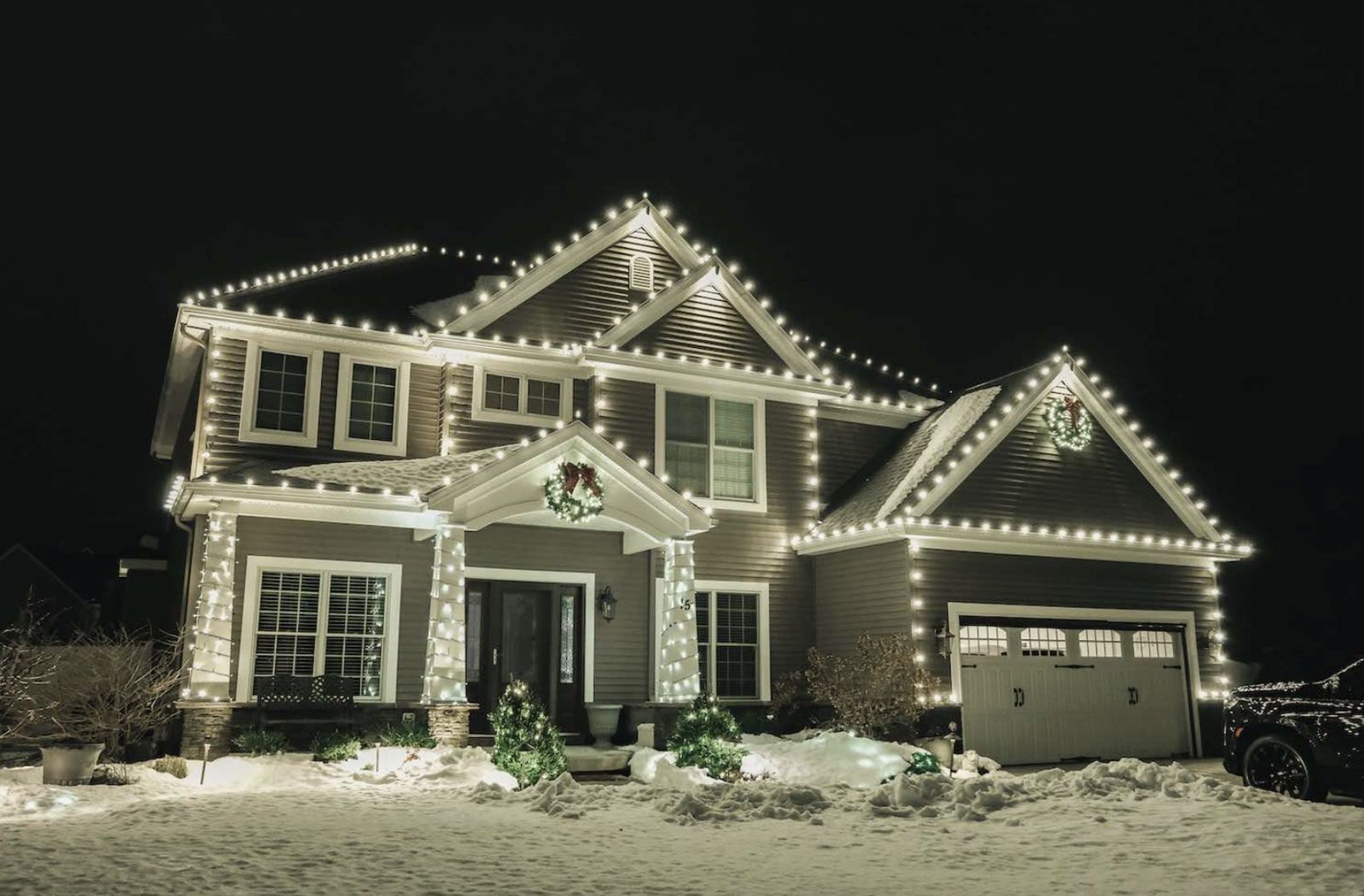 Two-story home with warm white Christmas lights along every roofline, wrapped porch columns and wreaths